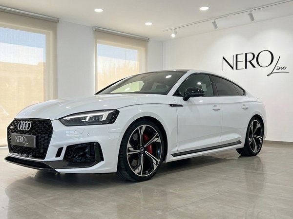 Audi RS5 — Luxusauto mieten in Alicante Der weiße Audi RS5 wird aus einer Dreiviertel-Frontansicht in einem modernen Showroom mit dem Nero Line Logo in Alicante präsentiert.