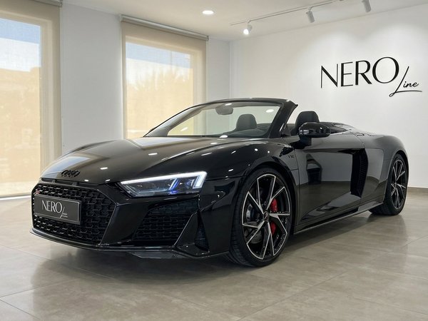 Audi R8 Spyder — Vista frontal tres cuartos en showroom Un Audi R8 Spyder descapotable negro brillante está aparcado en un luminoso concesionario, visto desde un ángulo frontal tres cuartos en Marbella.