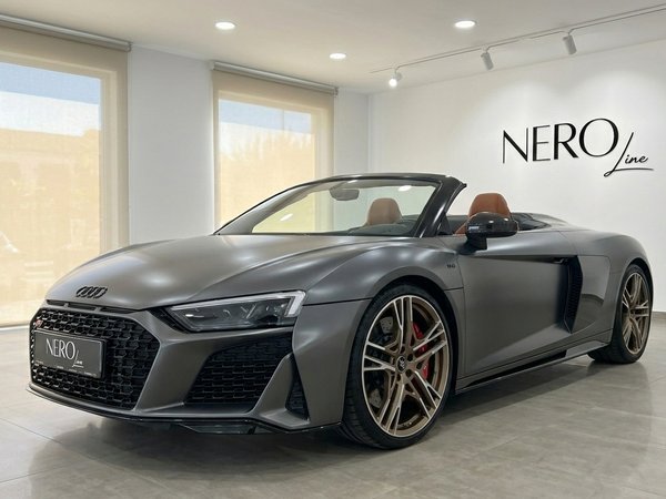 Audi R8 Spyder — Vista frontal tres cuartos El Audi R8 Spyder descapotable gris oscuro mate está aparcado en el interior, mostrando su vista frontal tres cuartos y lujoso interior marrón en Marbella.