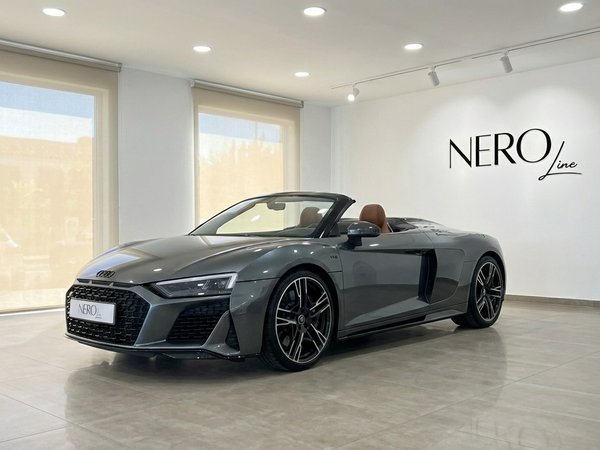 Audi R8 V10 Spyder Performance — Showroom Marbella The metallic gray 2023 Audi R8 V10 Spyder Performance convertible, with its top down and brown interior, is showcased in a showroom in Marbella.