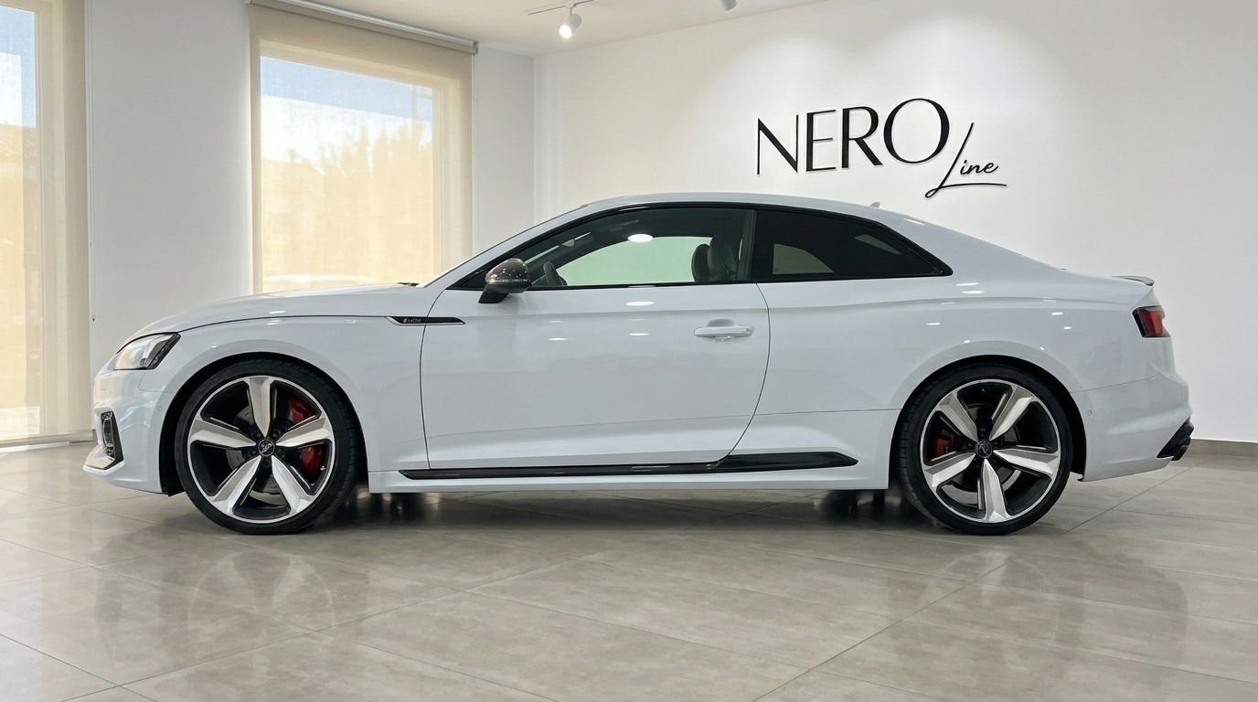 The white 2021 Audi RS5 is parked indoors, showcasing its sleek side profile and distinctive wheels in Alicante.