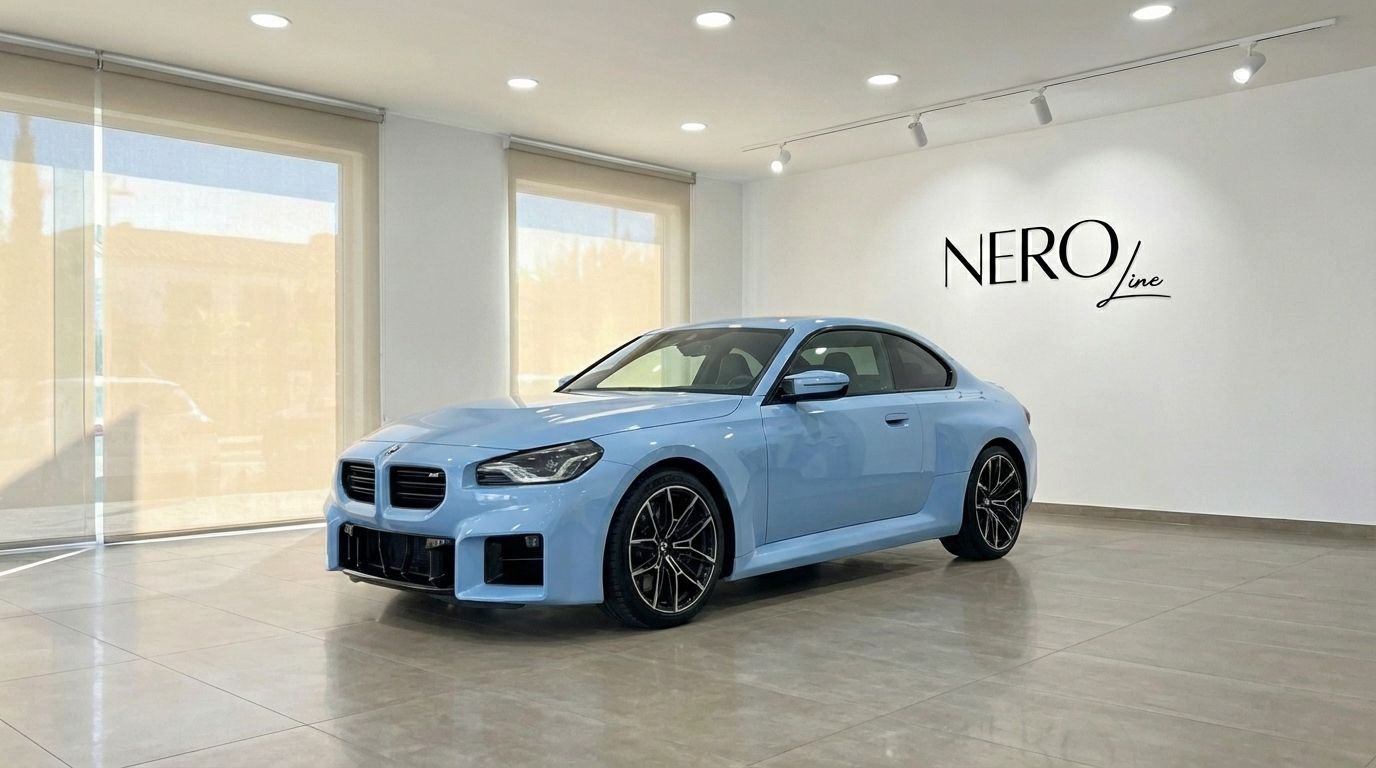 A light blue BMW M2 Competition, 2025 model, is parked in a showroom in Alicante, showing its front three-quarter view.
