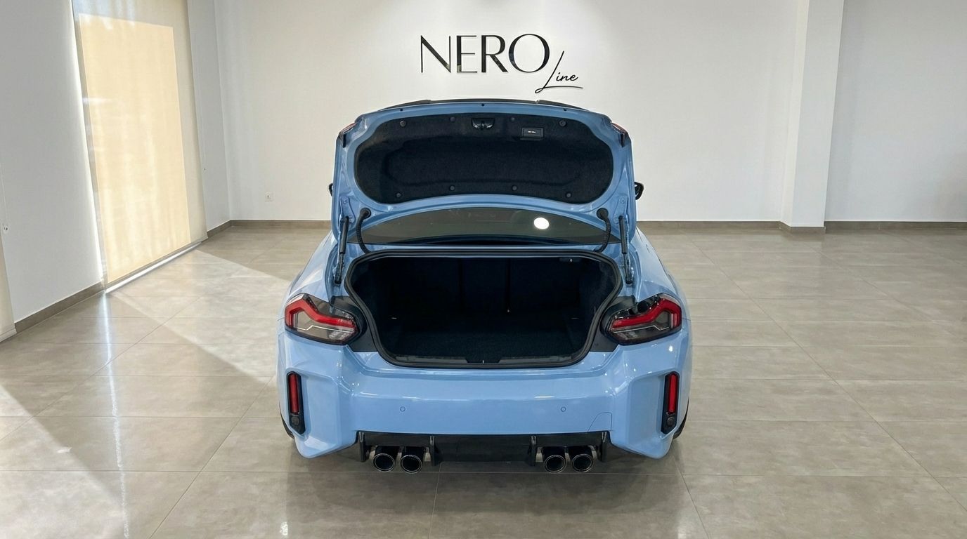 The light blue BMW M2 Competition is shown from the rear with its trunk open, parked in a showroom in Alicante.