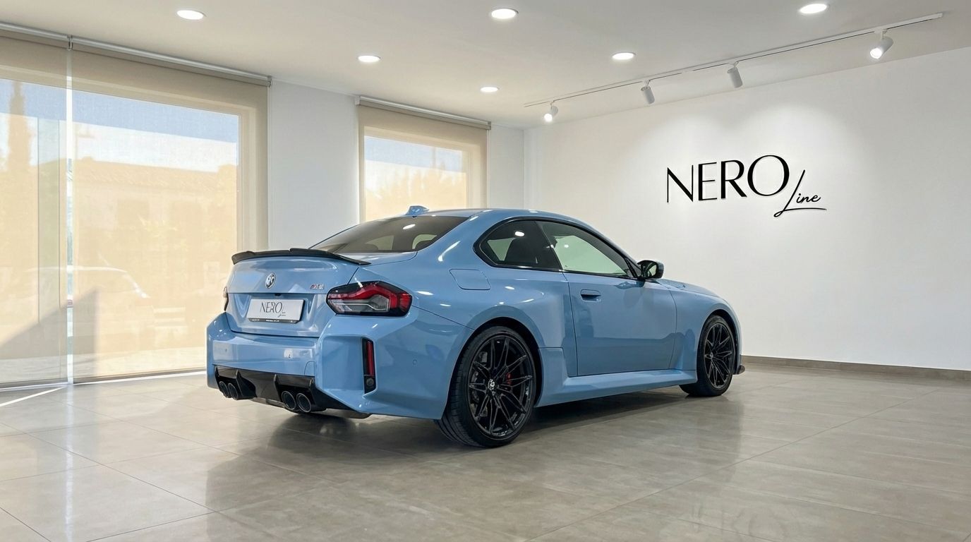 The light blue BMW M2 Competition is showcased from its dynamic rear three-quarter angle inside a modern showroom in Alicante.