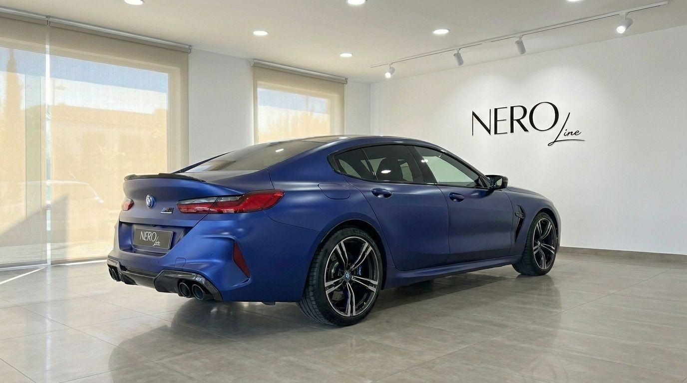 The matte blue BMW M8 Competition Gran Coupe is showcased from its rear three-quarter view inside a modern showroom in Marbella.
