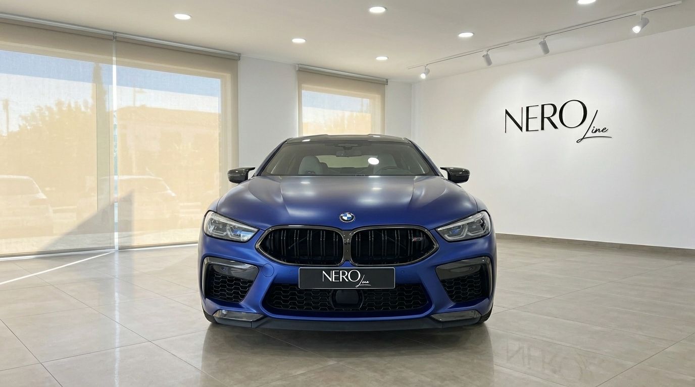 The matte blue BMW M8 Competition Gran Coupe is showcased from the front, parked inside a modern showroom in Marbella.
