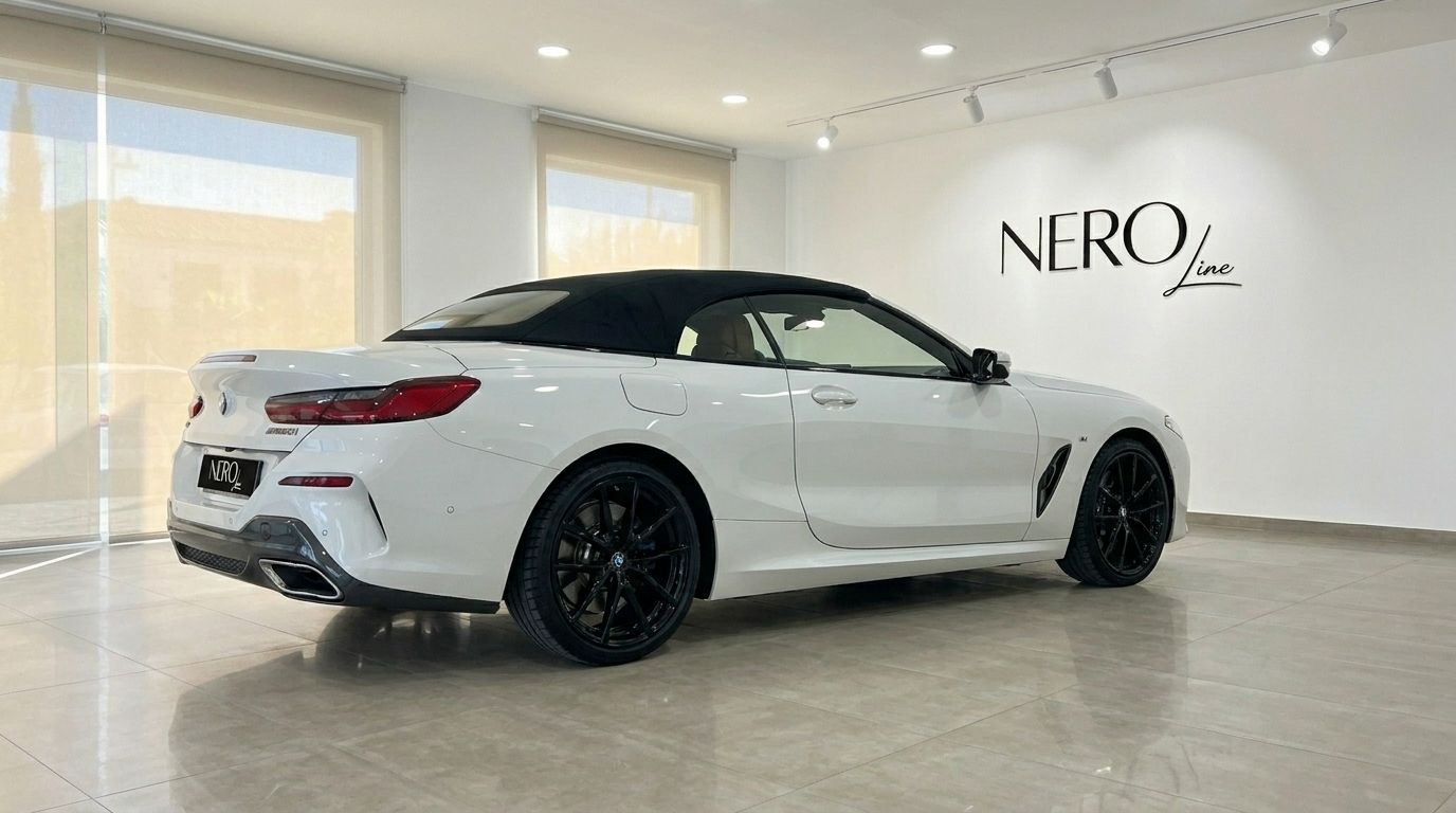 The white BMW M850i Cabriolet with its black soft top up is parked in a showroom in Alicante, seen from the rear three-quarter angle.