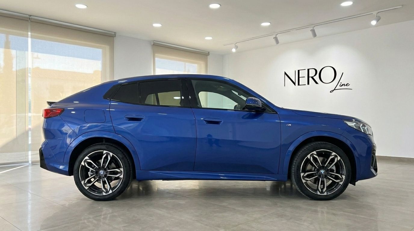The blue 2025 BMW X2 is shown in a side profile view, parked indoors at a showroom in Alicante.