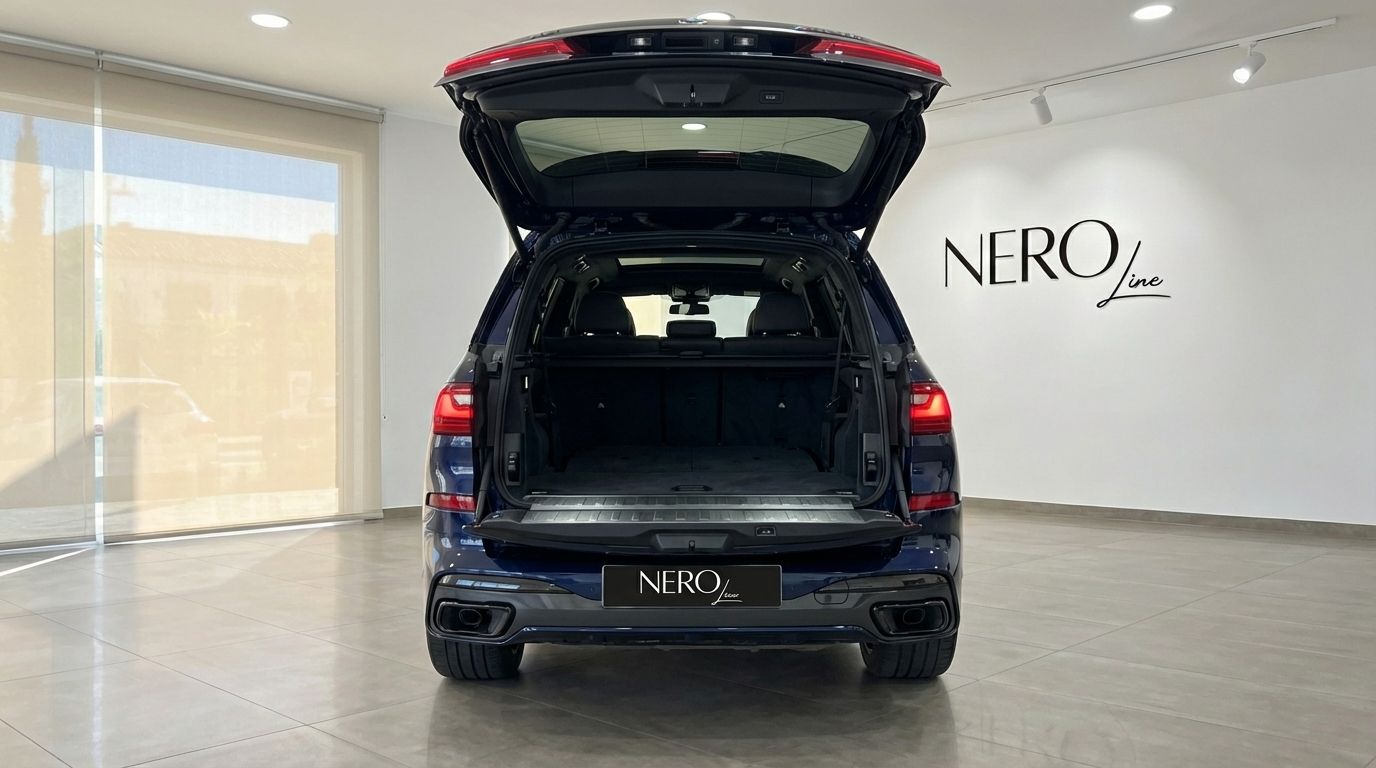 El BMW X7 M60i azul oscuro de 2024 se muestra desde la parte trasera con el maletero abierto, revelando su espaciosa área de carga en un concesionario de Madrid.