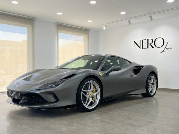 Ferrari F8 Coupe — Exhibición en showroom en Ibiza El Ferrari F8 Coupe gris mate se exhibe en un concesionario en Ibiza, mostrando su elegante vista frontal tres cuartos y pinzas de freno amarillas.