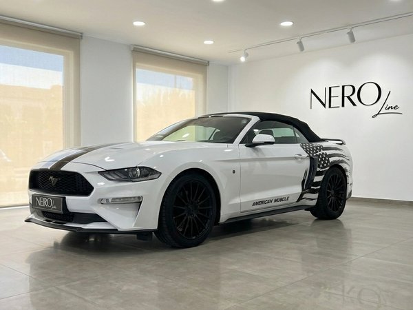 Ford Mustang 2.3 Convertible — White with American flag The white Ford Mustang 2.3 Convertible with black racing stripes and an American flag decal is parked indoors in Benidorm.