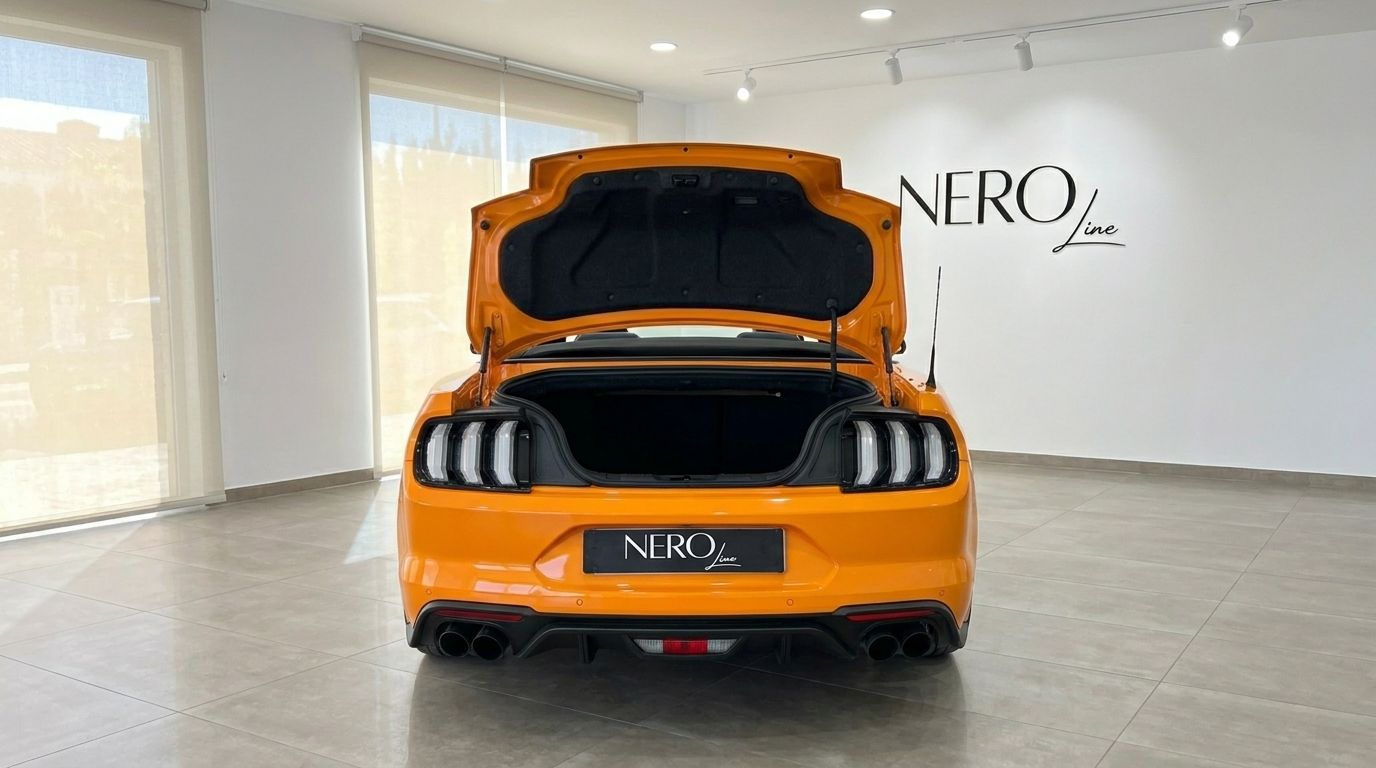 The rear view of an orange Ford Mustang 2.3 EcoBoost Convertible with its trunk open, parked in a showroom in Alicante.