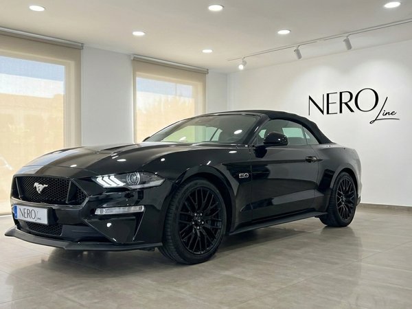 Ford Mustang GT 5.0 — Black Convertible, Alicante A sleek black Ford Mustang GT 5.0 convertible is displayed from a front three-quarter view in a bright Alicante showroom.