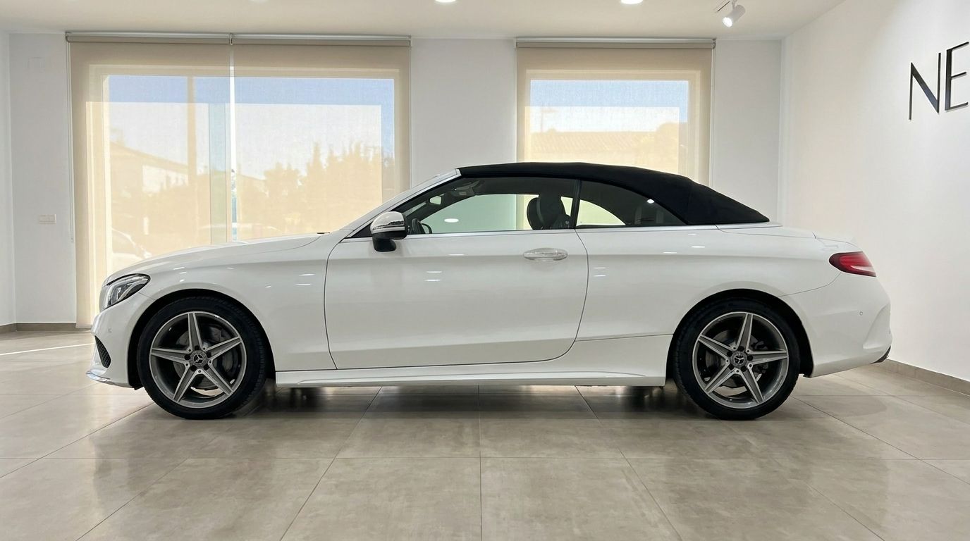 A white Mercedes-Benz C 200 Cabriolet with its black soft top up, shown in a side profile view inside a showroom in Alicante.