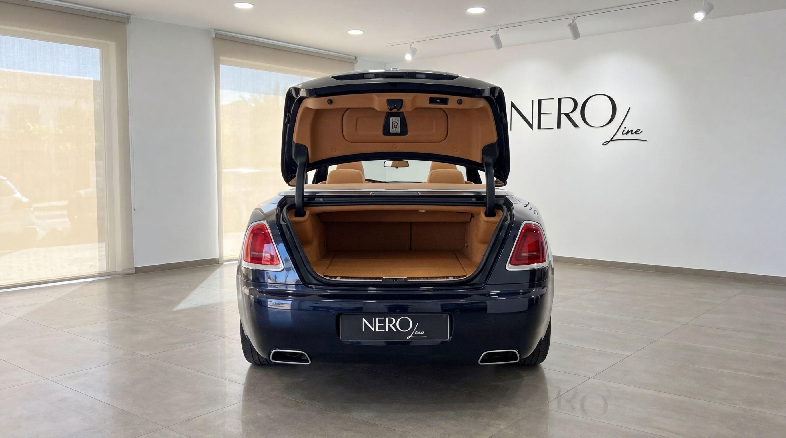 The midnight blue Rolls-Royce Dawn is shown from the rear with its trunk open, revealing the tan leather interior in a Barcelona showroom.