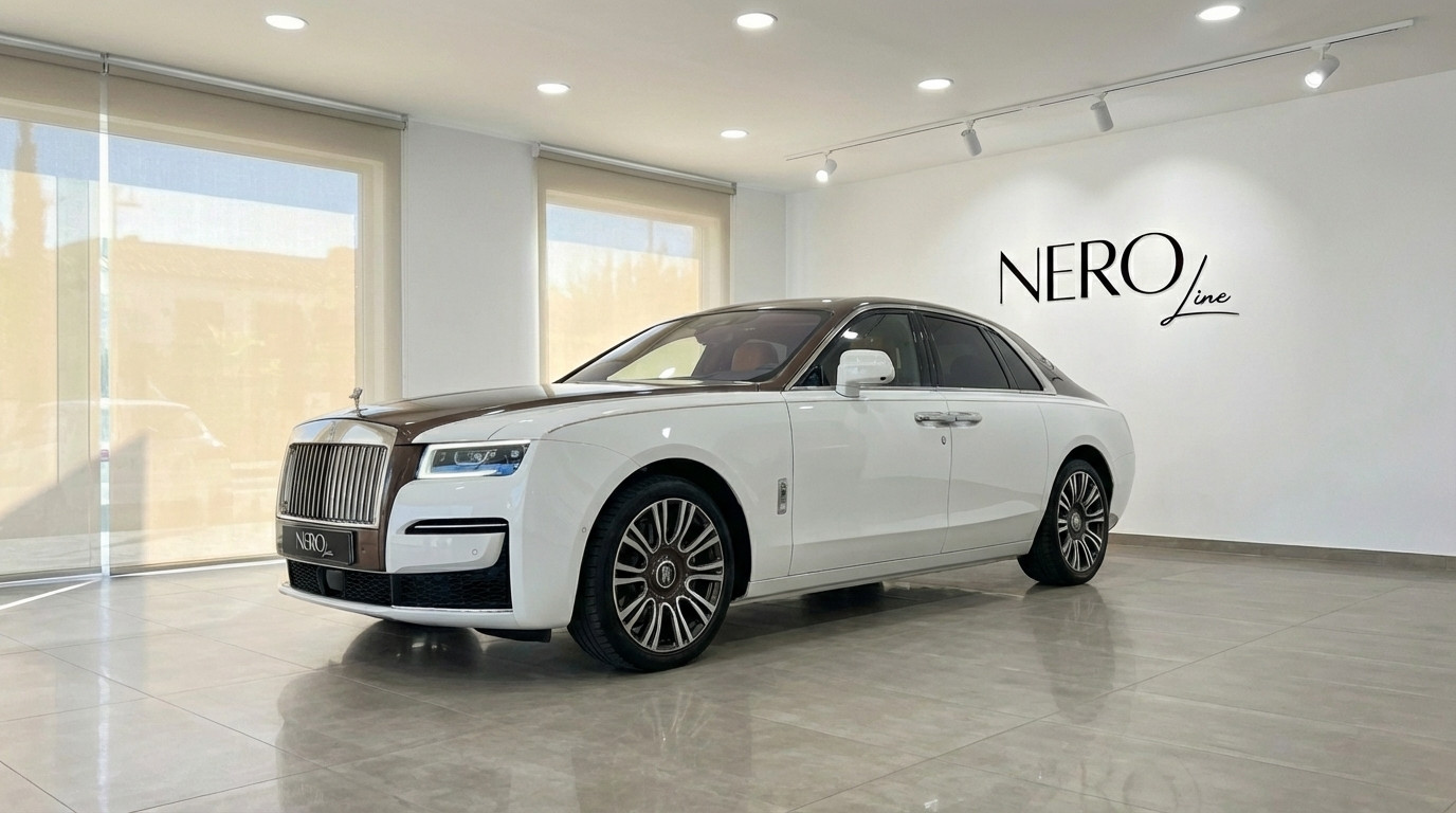 Rolls-Royce Ghost Amber Roads — Sedán de Lujo Madrid El sedán de lujo Rolls-Royce Ghost Amber Roads blanco y marrón 2024 está aparcado en el interior en Madrid, visto desde un ángulo frontal de tres cuartos.