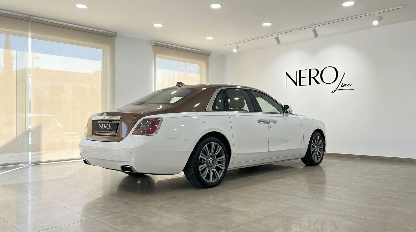 El sedán de lujo Rolls-Royce Ghost Amber Roads 2024 blanco y marrón está aparcado en un concesionario en Madrid, visto desde el ángulo trasero tres cuartos.