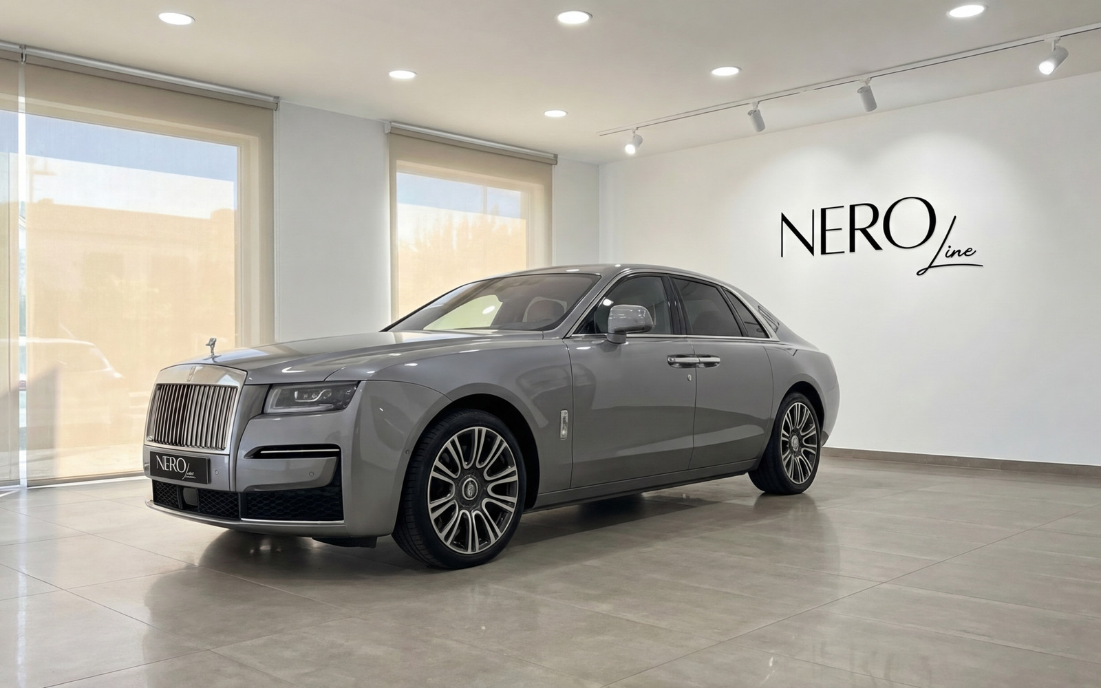 Rolls-Royce Ghost — Vue en showroom à Madrid Une Rolls-Royce Ghost gris métallisé, présentant son élégant profil avant et latéral dans un showroom moderne à Madrid.