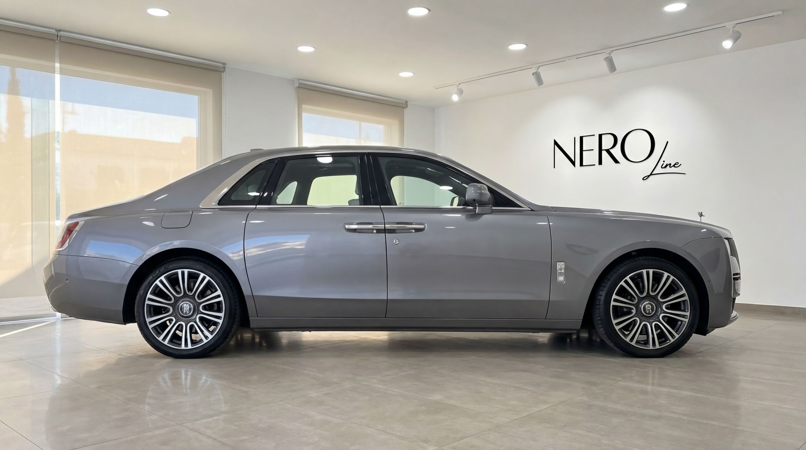 The elegant metallic gray Rolls-Royce Ghost luxury sedan is showcased in a side profile view within a well-lit showroom in Valencia.