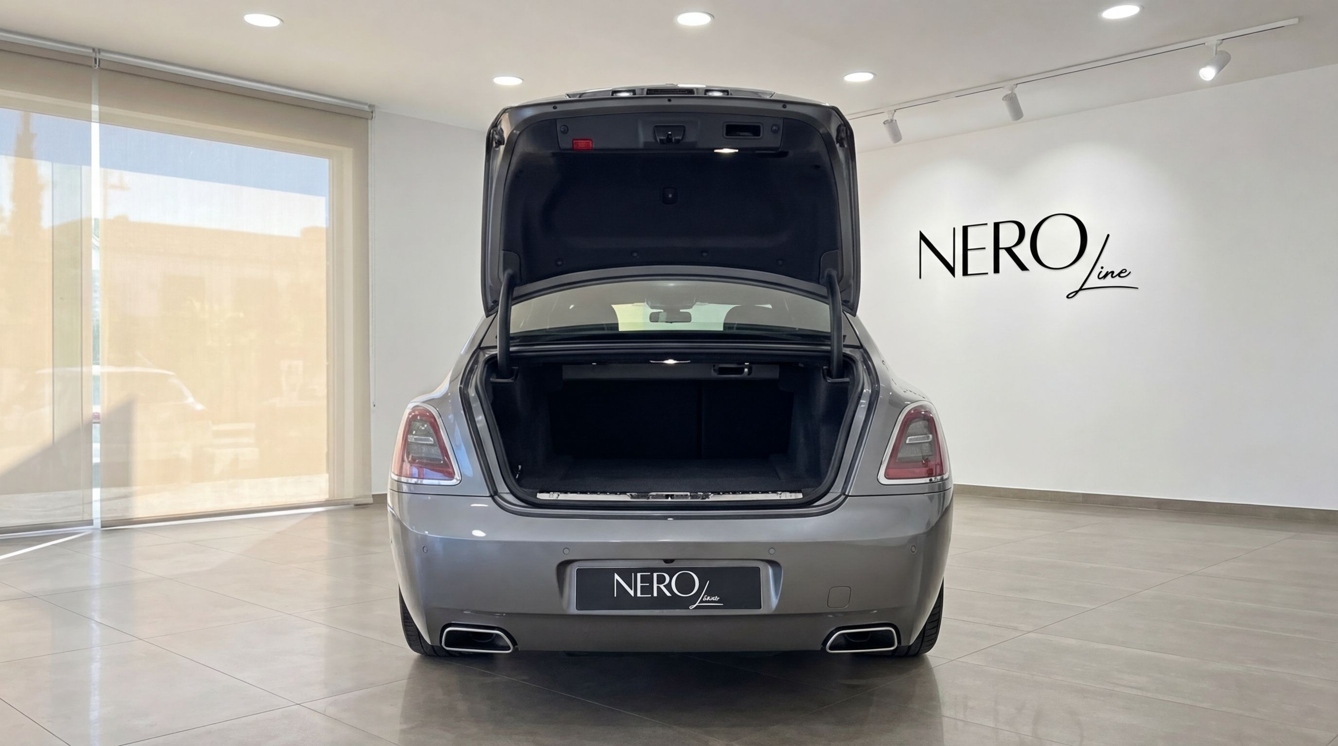 The metallic gray Rolls-Royce Ghost is shown from the rear with its trunk open, revealing ample storage space in a Valencia showroom.